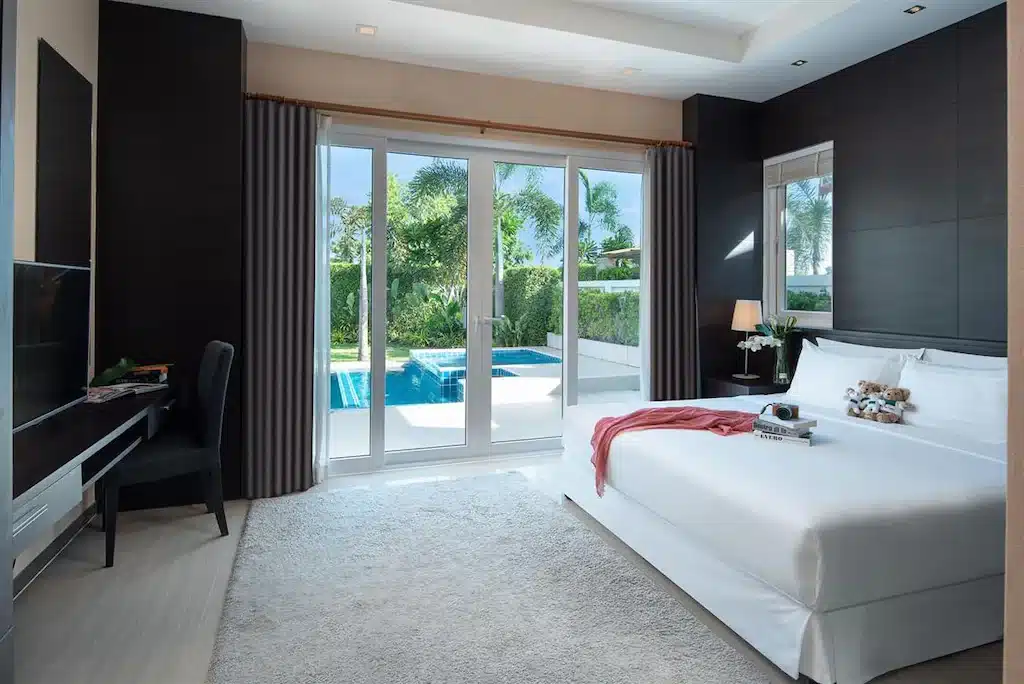 ห้องนอนสมัยใหม่พร้อมเตียงขนาดใหญ่ โต๊ะทำงาน และประตูกระจกที่เปิดออกสู่สระว่ายน้ำส่วนตัวและสวน—เหมาะสำหรับการพักผ่อนในที่พักติดทะเลพัทยาที่มีสไตล์ เพียงไม่กี่ก้าวจากชายหาด.