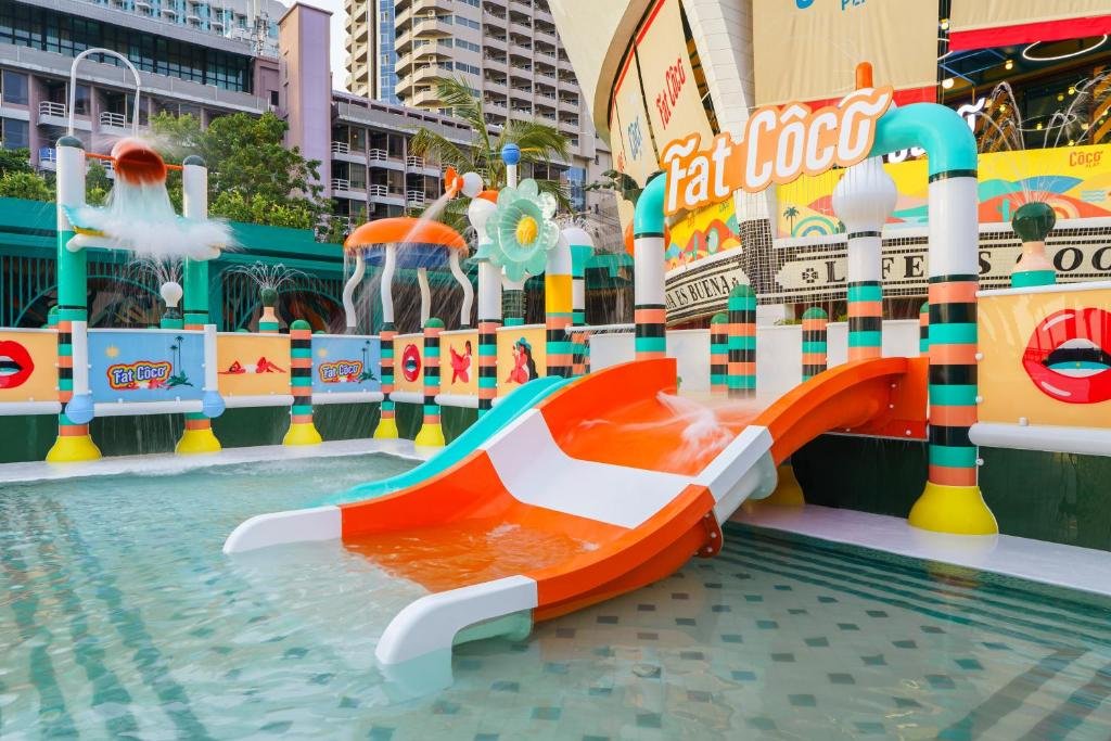 สนามเด็กเล่นน้ำสีสันสดใสที่ที่พักติดทะเลพัทยา มีสไลเดอร์สีส้มและขาวสองตัว น้ำตื้น ตกแต่งอย่างสนุกสนาน และมีป้าย "Fat Coco" อยู่ด้านหลัง.