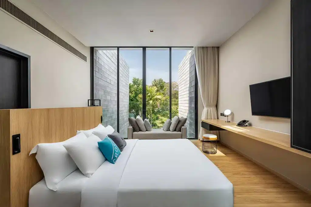 ห้องพักโรงแรมสมัยใหม่พร้อมเตียงขนาดใหญ่ ทีวีติดผนัง โต๊ะไม้ และหน้าต่างบานใหญ่จากพื้นจรดเพดานที่มองเห็นต้นไม้และที่นั่งริมหน้าต่างที่แสนสบาย—ทางเลือกที่เหมาะสำหรับทุกคนที่กำลังมองหา โรงแรมพัทยาติดทะเล สำหรับการพักผ่อนที่ผ่อนคลาย.