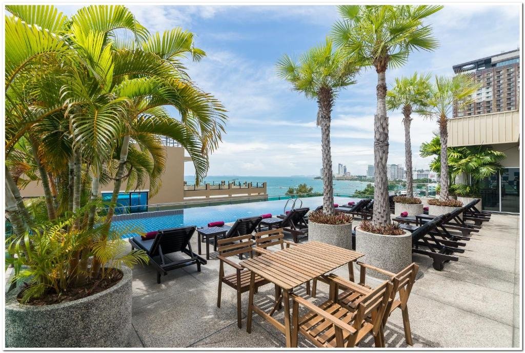 บริเวณสระว่ายน้ำกลางแจ้งพร้อมต้นปาล์ม โต๊ะไม้ เก้าอี้อาบแดด และวิวทะเลที่สวยงาม—เหมาะสำหรับการผ่อนคลายที่ ที่พักติดทะเลพัทยา พร้อมตึกในเมืองไกลออกไปภายใต้ท้องฟ้าที่มีเมฆบางส่วน.