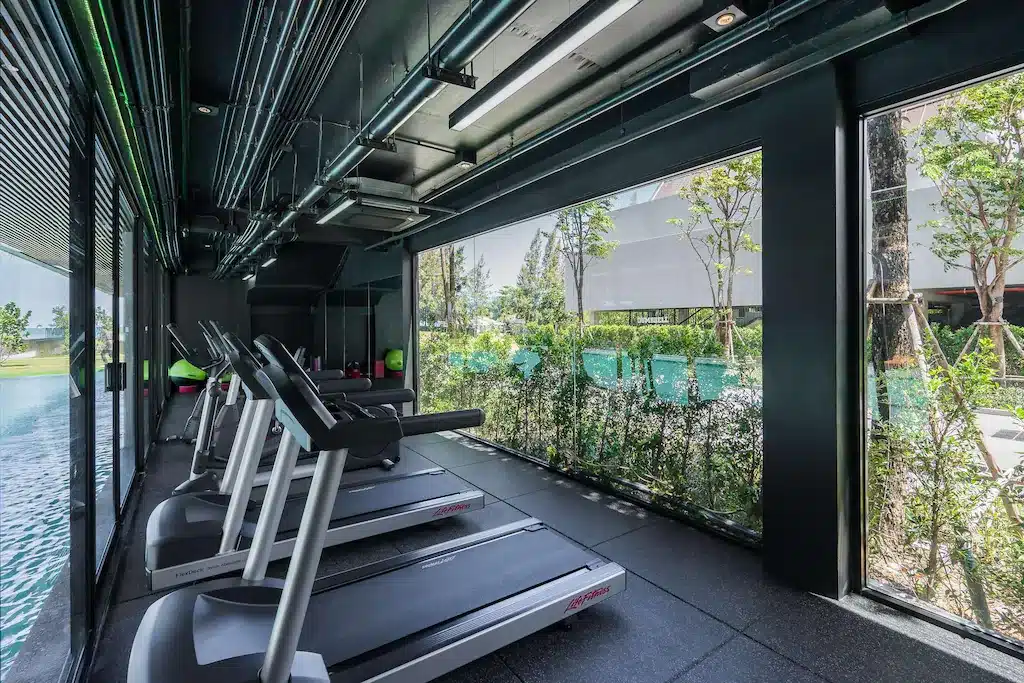 ลู่วิ่งสี่เครื่องเรียงกันอยู่ในยิมทันสมัยที่มีหน้าต่างบานใหญ่มองเห็นพื้นที่สีเขียวและสระว่ายน้ำภายนอกของโรงแรมติดทะเลพัทยา เพื่อประสบการณ์การออกกำลังกายที่สดชื่นริมทะเล.