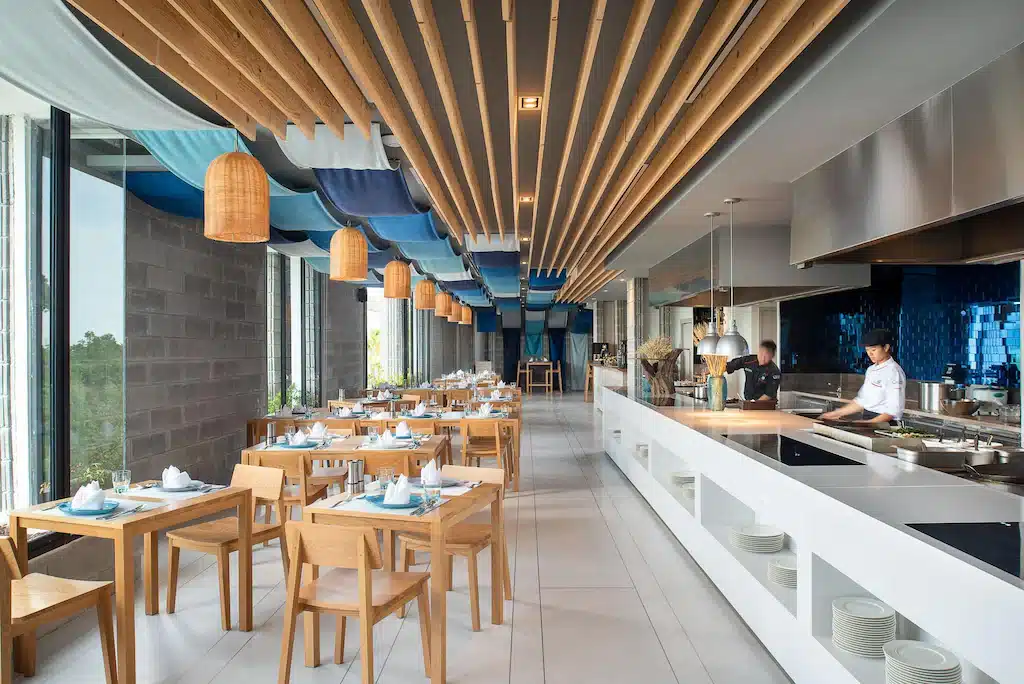 ร้านอาหารสมัยใหม่ตกแต่งภายในด้วยโต๊ะและเก้าอี้ไม้ ครัวเปิดที่เชฟกำลังปรุงอาหาร หน้าต่างบานใหญ่ และเพดานประดับด้วยคานไม้และผ้าสีน้ำเงิน ตั้งอยู่ในโรงแรมติดทะเลพัทยา มอบประสบการณ์การรับประทานอาหารริมชายหาดที่น่าประทับใจ.