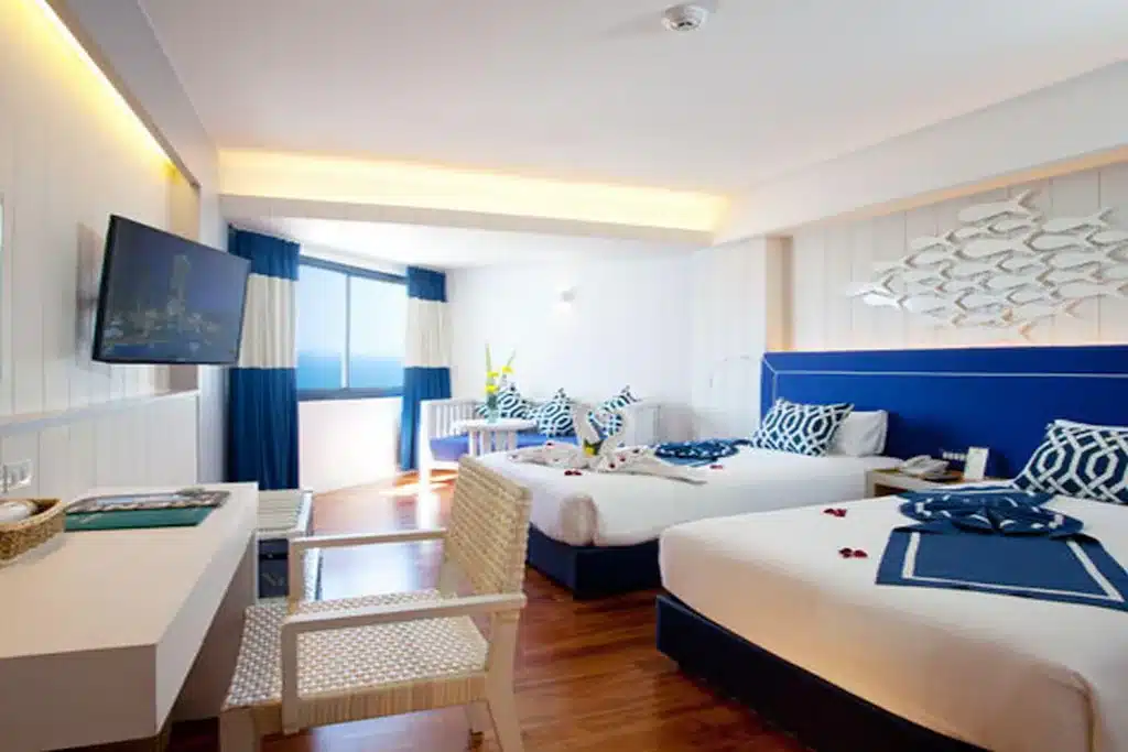 ห้องพักโรงแรมสว่างสดใสพร้อมเตียงคู่สองเตียง ตกแต่งด้วยสีน้ำเงินและสีขาว ทีวีติดผนัง หน้าต่างวิวทะเล และเฟอร์นิเจอร์ทันสมัย—เหมาะสำหรับผู้ที่มองหา โรงแรมติดทะเลพัทยา หรือ ที่พักติดทะเลพัทยา ที่มีความสะดวกสบายและสไตล์.