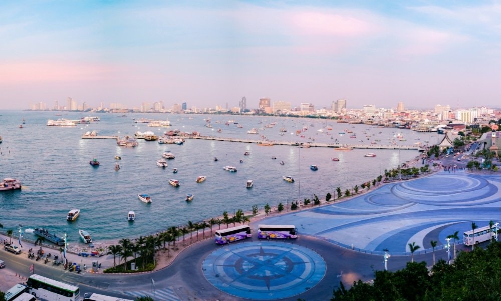 ภาพพาโนรามาของเมืองชายฝั่งที่มีเรือลอยอยู่ในน้ำ ชายฝั่งโค้งที่เรียงรายไปด้วยตึกสูงสมัยใหม่และโรงแรมติดทะเลพัทยา พร้อมทางเดินกว้างที่มีลวดลายสวยงามใต้ท้องฟ้าสีพาสเทล.