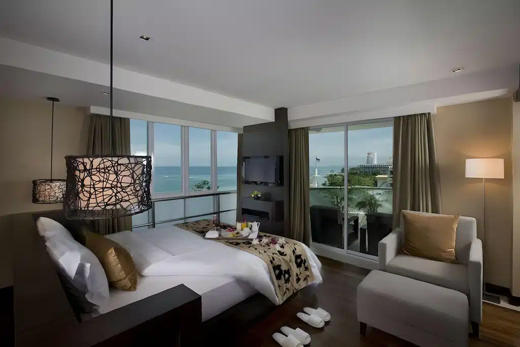 ห้องพักริมทะเลพัทยาสมัยใหม่พร้อมเตียง, เก้าอี้นวม, รองเท้าแตะ, และถาดอาหารเช้า, มีหน้าต่างขนาดใหญ่และระเบียงพร้อมวิวทะเลและตึกในเมือง.