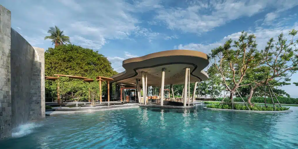 สระว่ายน้ำกลางแจ้งพร้อมศาลาหลังคาโค้งทันสมัย ณ ที่พักพัทยาติดทะเล รายล้อมด้วยต้นไม้และท้องฟ้าสีครามใส.