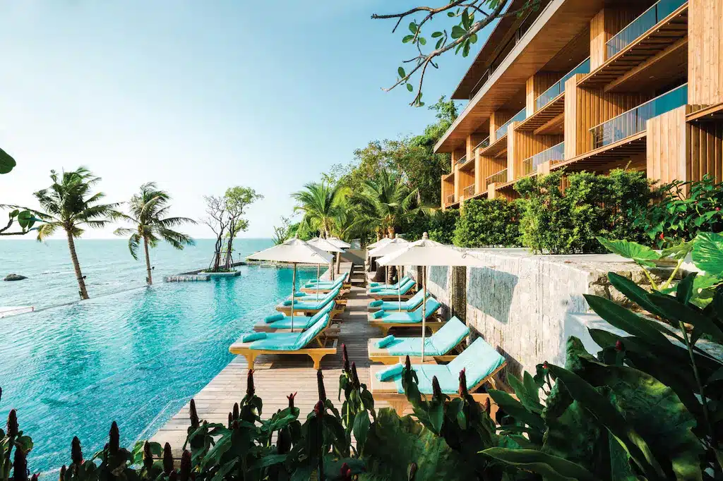 สระว่ายน้ำไร้ขอบพร้อมเก้าอี้อาบแดดสีฟ้าเทอร์ควอยซ์และร่มสีขาวตั้งอยู่ข้างโรงแรมไม้สมัยใหม่; ต้นปาล์มและวิวทะเลภายใต้ท้องฟ้าแจ่มใส ณ ที่พักพัทยาติดทะเลสุดหรูสำหรับการพักผ่อนริมทะเลที่สมบูรณ์แบบ.