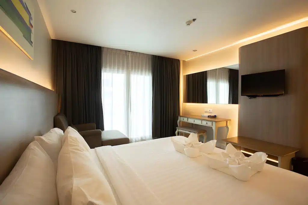ห้องพักโรงแรมสมัยใหม่พร้อมเตียงขนาดใหญ่ ผ้าปูที่นอนสีขาว โทรทัศน์ติดผนัง โต๊ะทำงาน เก้าอี้นวม และหน้าต่างบานใหญ่พร้อมผ้าม่าน—เหมาะสำหรับการพักผ่อนที่โรงแรมติดทะเลพัทยา พร้อมการจัดวางผ้าขนหนูตกแต่งอย่างสวยงามบนเตียง.