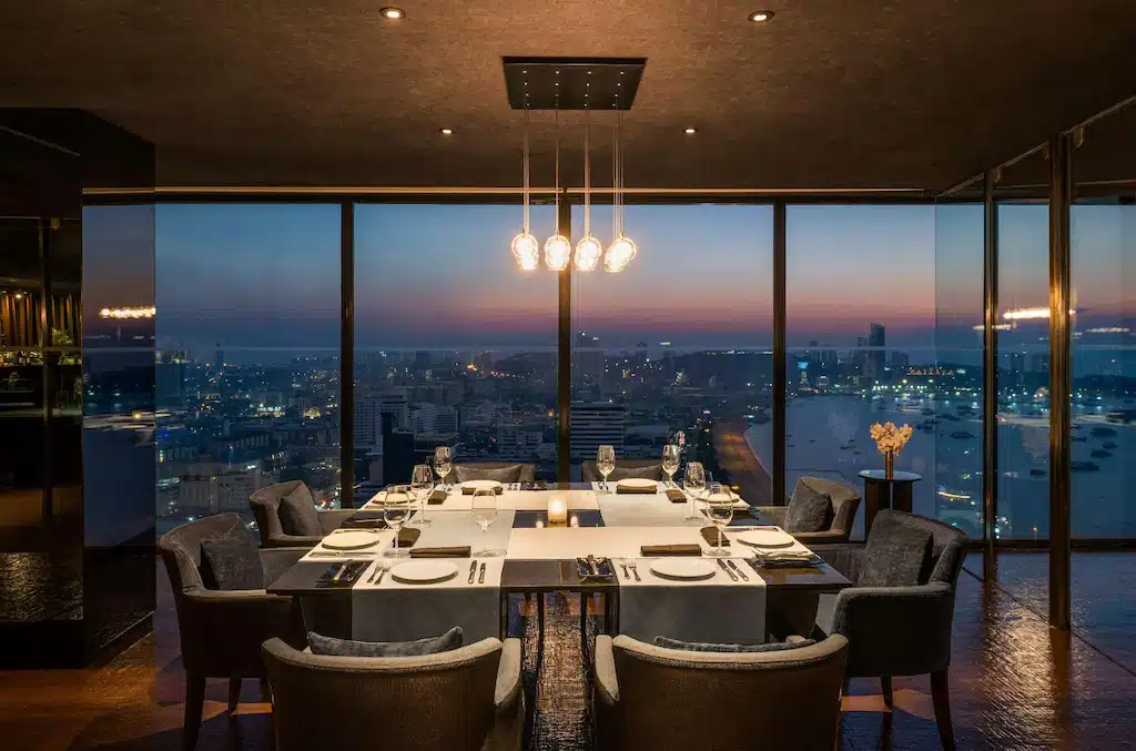 ชุดโต๊ะอาหารหรูหราสำหรับ 6 ท่าน ในภัตตาคารสไตล์โมเดิร์น พร้อมหน้าต่างกระจกบานใหญ่จากพื้นจรดเพดาน มองเห็นทัศนียภาพของเมืองในยามพระอาทิตย์ตกดิน เหมาะสำหรับแขกที่ต้องการสัมผัสประสบการณ์การรับประทานอาหารที่ไม่อาจลืมได้ที่โรงแรมพัทยาติดทะเล.