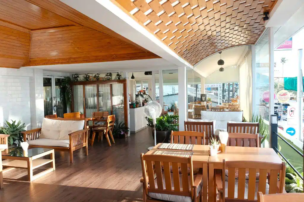 ภายในคาเฟ่ที่สว่างและทันสมัย ตกแต่งด้วยเฟอร์นิเจอร์ไม้ หน้าต่างบานใหญ่ และต้นไม้ในกระถาง—เหมาะสำหรับการพักผ่อนหลังจากสำรวจ โรงแรมติดทะเลพัทยา พร้อมโซฟาและโต๊ะรับประทานอาหารที่จัดวางบนพื้นไม้ขัดเงา.