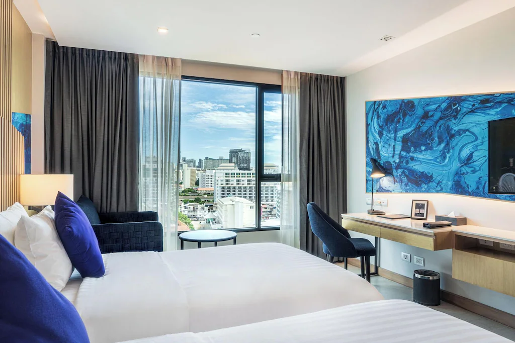 ห้องพักโรงแรมสมัยใหม่พร้อมเตียงสองเตียง ตกแต่งด้วยสีน้ำเงิน หน้าต่างบานใหญ่พร้อมวิวเมือง โต๊ะทำงาน เก้าอี้ งานศิลปะบนผนัง และทีวีติดผนัง—เหมาะสำหรับผู้ที่มองหาโรงแรมพัทยาติดทะเลที่มีสไตล์สำหรับการพักผ่อนริมชายหาด.