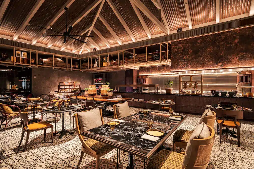 ร้านอาหารหรูหราภายในตกแต่งด้วยพื้นกระเบื้องลาย, โต๊ะหินอ่อน, เก้าอี้บุนวม, และครัวเปิด—เหมาะสำหรับแขกที่ต้องการประสบการณ์การรับประทานอาหารที่หรูหราในโรงแรมติดทะเลพัทยาใต้ภายใต้เพดานโค้งสูงพร้อมแสงสว่างอบอุ่น.