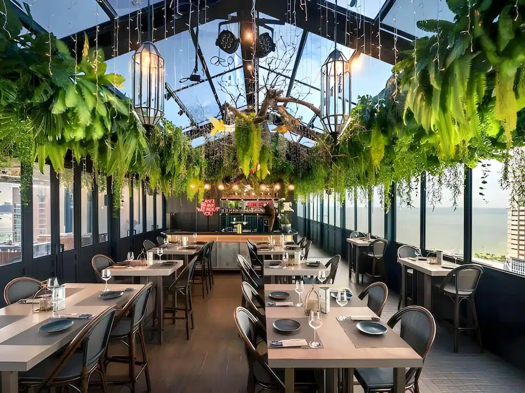 ร้านอาหารบนดาดฟ้าสมัยใหม่พร้อมหน้าต่างบานใหญ่ โต๊ะไม้ เก้าอี้สีดำ ต้นไม้แขวน และไฟประดับสาย มองเห็นวิวทะเล เหมาะสำหรับแขกของที่พักพัทยาติดทะเลหรือโรงแรมติดทะเลพัทยาที่ต้องการประสบการณ์การรับประทานอาหารที่มีสไตล์.