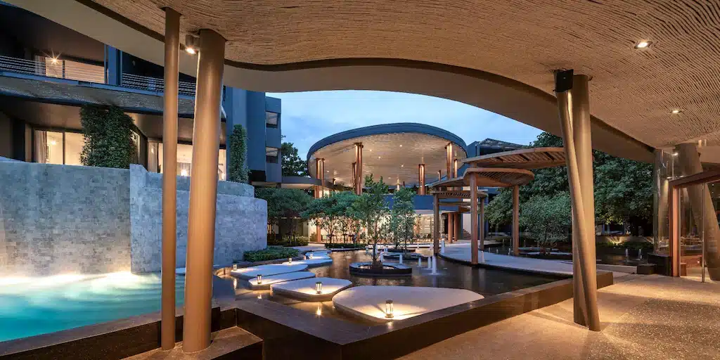 ลานโรงแรมสมัยใหม่พร้อมสระว่ายน้ำ, กระถางต้นไม้ทรงกลม, น้ำพุ, และสถาปัตยกรรมสมัยใหม่—โรงแรมพัทยาติดทะเลแห่งนี้มอบทางเดินที่มีหลังคาคลุมพร้อมแสงไฟอบอุ่นในยามเย็นเพื่อการพักผ่อนริมทะเลที่เงียบสงบ.