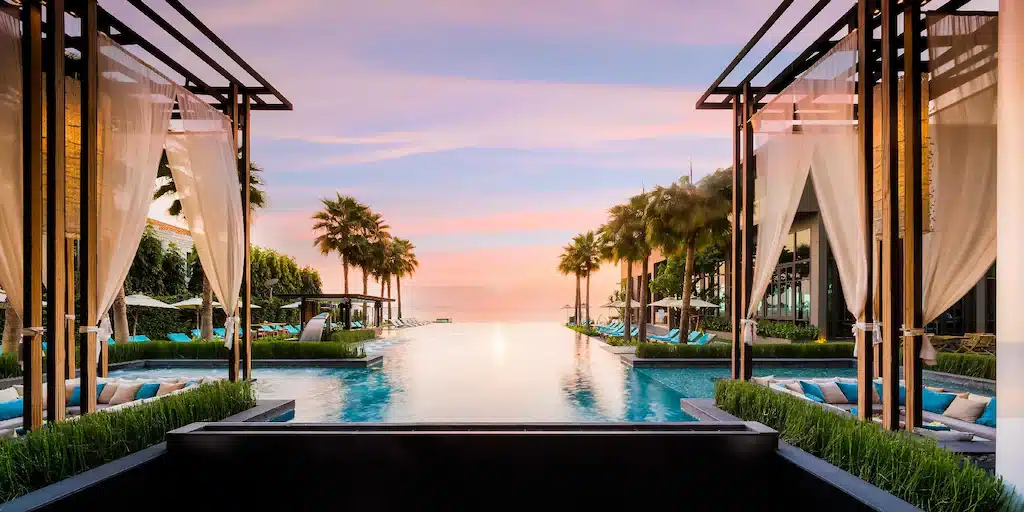 สระว่ายน้ำแบบอินฟินิตี้ล้อมรอบด้วยต้นปาล์มและศาลาพักผ่อน ณ รีสอร์ทติดทะเลพัทยา พร้อมวิวท้องฟ้ายามพระอาทิตย์ตกดินอันงดงามเป็นฉากหลัง.