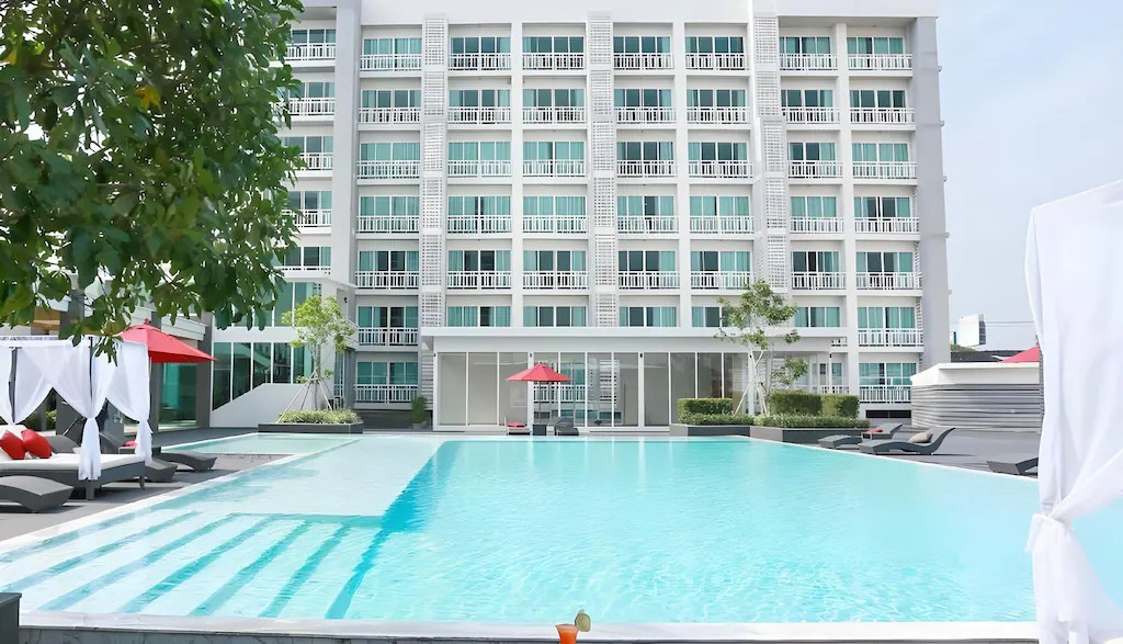 โรงแรมพัทยาติดทะเลสไตล์โมเดิร์นพร้อมระเบียงมองเห็นสระว่ายน้ำกลางแจ้งขนาดใหญ่ ล้อมรอบด้วยเก้าอี้อาบแดด ศาลาสีขาว และร่มสีแดง—เหมาะ ที่พักระยอง สำหรับการพักผ่อนริมชายหาดอย่างแท้จริง.