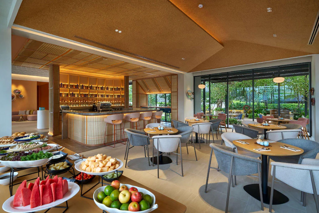 ร้านอาหารสมัยใหม่ที่มีการจัดโต๊ะอย่างเป็นระเบียบ มีบาร์กลาง และบุฟเฟต์ผล ที่พักระยอง ไม้สดและสลัด หน้าต่างบานใหญ่เปิดรับวิวสีเขียว สร้างบรรยากาศที่เชิญชวนภายในโรงแรมติดทะเลพัทยาแห่งนี้ ให้แขกได้ผ่อนคลายริมทะเล.