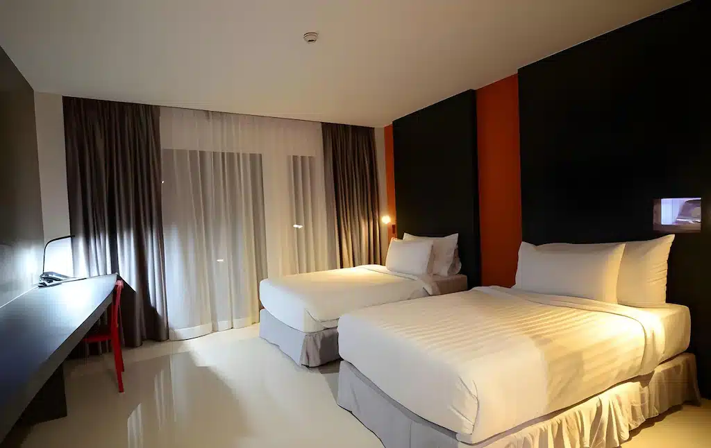 ห้องพักโรงแรมสมัยใหม่พร้อมเตียงเดี่ยวสองเตียง ชุดเครื่องนอนสีขาว   โรงแรมระยอง ผนังเน้นสีเข้ม โต๊ะพร้อมโคมไฟ และหน้าต่างบานใหญ่จากพื้นจรดเพดานพร้อมผ้าม่านโปร่งและผ้าม่านกันแสง—เหมาะสำหรับการเข้าพักของคุณที่ โรงแรมพัทยาติดทะเล.