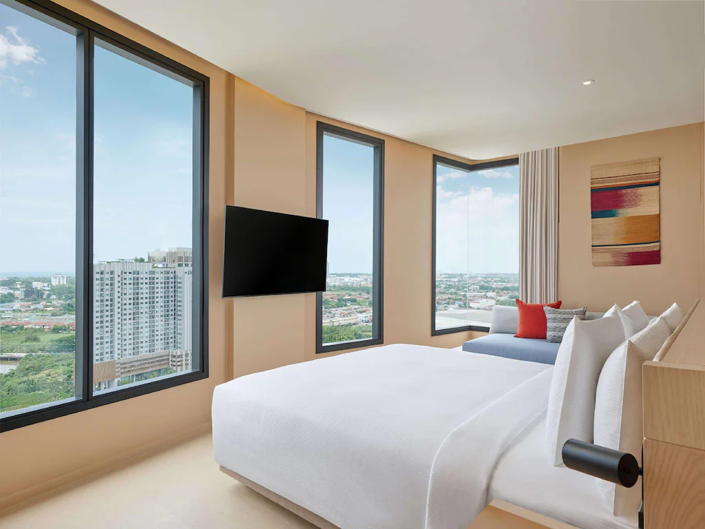 ห้องพักโรงแรมสมัยใหม่ที่ โรงแรม ที่พักระยอง พัทยาติดทะเล, พร้อมหน้าต่างบานใหญ่จากพื้นจรดเพดาน, เตียงผ้าลินินสีขาว, โซฟา, โทรทัศน์ติดผนัง, และวิวเมืองที่สวยงามในเวลากลางวัน.