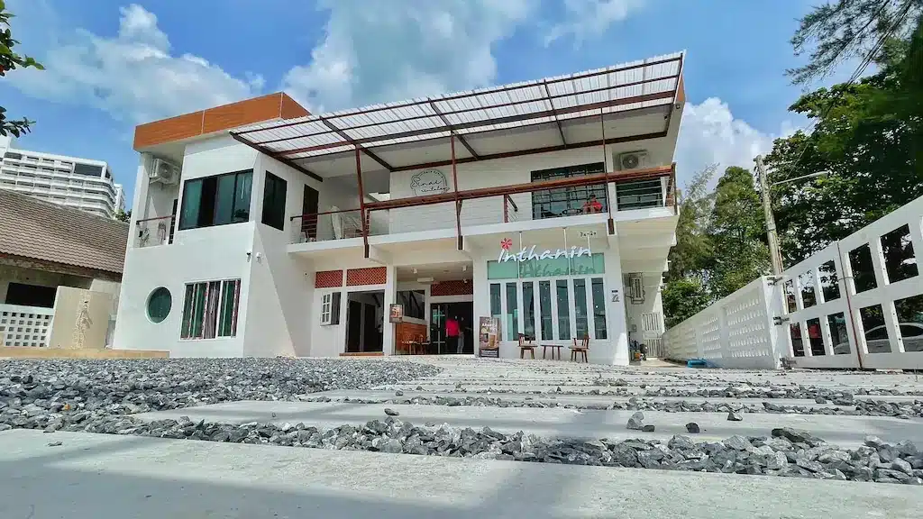 อาคารสองชั้นสีขาวพร้อมระเบียงที่มีหลังคาคลุมและป้าย "Indahcafe" ที่ทางเข้า; ทางเดินกรวดนำไปสู่ประตูหน้า เหมาะสำหรับผู้ที่มองหาที่พักติดรวม ที่พักระยอง คัดมาแล้ว ติดทะเล วิวสวย พักผ่อนชิลใกล้กรุงเทพ โรงแรมระยอง บรรยากาศริมชายหาด ไม่ว่าจะเป็น รีสอร์ทระยอง โซนที่ติดทะเลหรือโซนในเมือง!!
ทะเลพัทยาที่อบอุ่น มีต้นไม้และอาคารอีกหลังอยู่ใกล้เคียง.