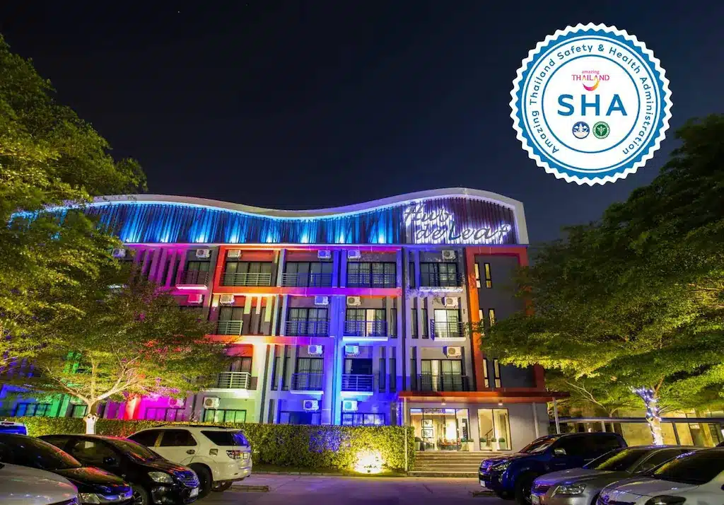 โรงแรมพัทยาติดทะเลสมัยใหม่หลายชั้นที่ประดับด้วยไฟหลากสีในยามค่ำคืน มี  โรงแรมระยอง รถยนต์จอดอยู่ด้านหน้า พร้อมป้ายรับรองความปลอดภัย "SHA" อยู่ที่มุมขวาบน.