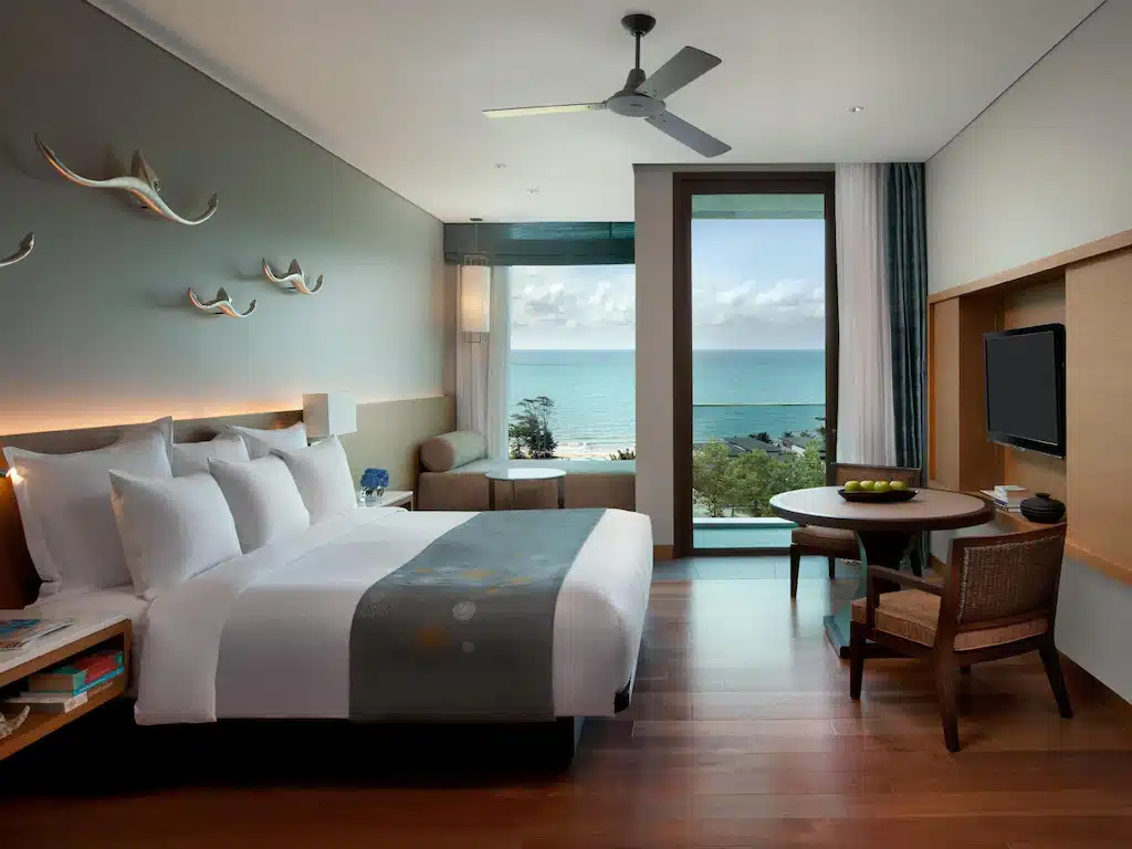 ห้องพักโรงแรมสมัยใหม่พร้อมเตียง  โรงแรมระยอง ขนาดใหญ่ พื้นที่นั่งเล่น โต๊ะพร้อมเก้าอี้ ทีวีติดผนัง และระเบียงที่มองเห็นวิวทะเล—เหมาะสำหรับผู้ที่มองหา โรงแรมติดทะเลพัทยา และการพักผ่อนริมทะเลที่ผ่อนคลาย.