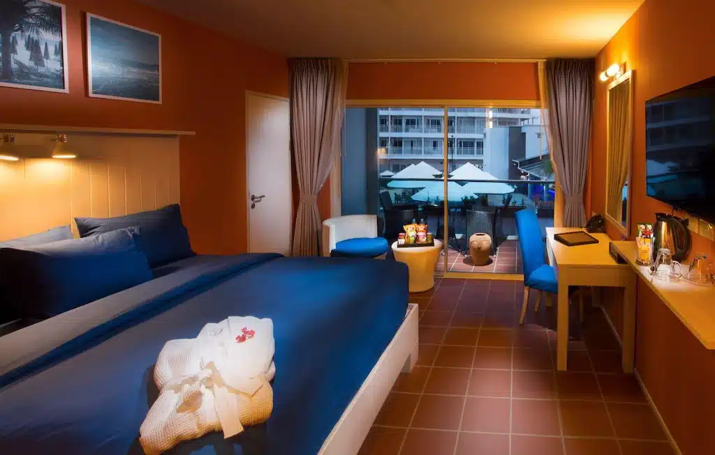โรงแรมติดทะเลพัทยา ห้องพักมีเตียง  โรงแรมระยอง ใหญ่ ผ้าปูที่นอนสีน้ำเงิน โต๊ะทำงาน เก้าอี้ และทีวี ระเบียงชมวิวสระว่ายน้ำ ร่มสีสดใส และอาคารรายล้อม เหมาะสำหรับการพักผ่อนริมชายหาด.