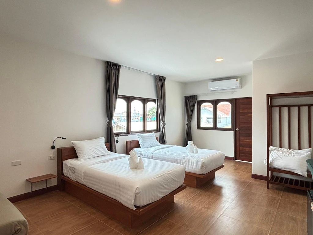 ห้องพักโรงแรมที่สว่างไสวที่  โรงแรมระยอง  ประกอบด้วยเตียงเดี่ยวสองเตียงพร้อมผ้าปูที่นอนสีขาว เตียงสองชั้น ประตูไม้ และหน้าต่างบานใหญ่พร้อมผ้าม่านสีเข้ม.