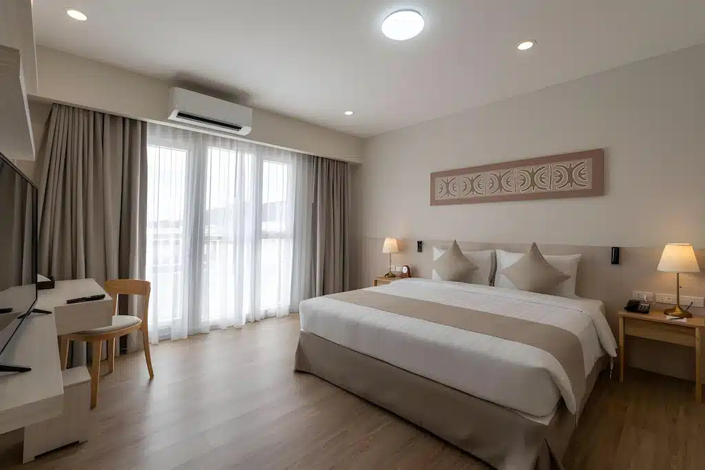 ห้องพักโรงแรมสมัยใหม่พร้อมเตียง  โรงแรมระยอง ขนาดใหญ่ โต๊ะข้างเตียงพร้อมโคมไฟ โต๊ะทำงานและเก้าอี้ โทรทัศน์ติดผนัง และหน้าต่างบานใหญ่จากพื้นจรดเพดานพร้อมผ้าม่านสีเบจ—เหมาะสำหรับการเข้าพักของคุณที่ โรงแรมติดทะเลพัทยา.
