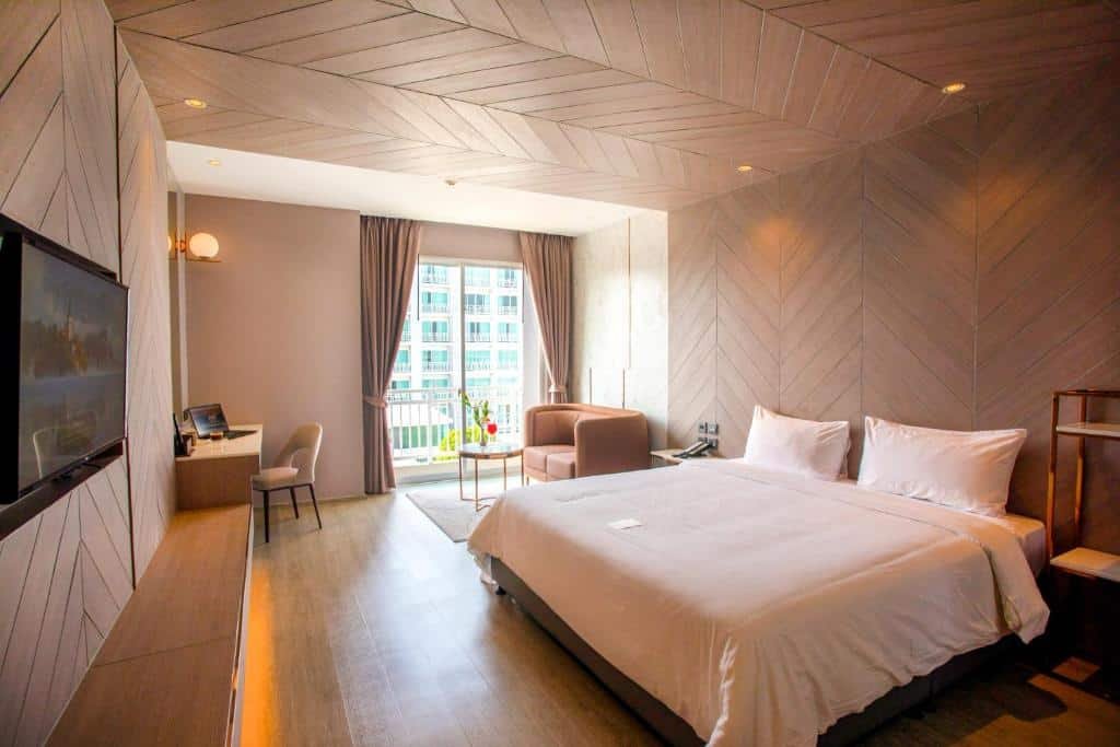 ห้องพักโรงแรมพร้อมเตียงขนาดใหญ่ โต๊ะทำงาน โทรทัศน์ เก้าอี้โซฟา และหน้าต่างพร้อมผ้าม่านที่มองเห็นวิว ที่พักระยอง เมือง เพลิดเพลินกับการตกแต่งด้วยไม้สมัยใหม่และแสงไฟอบอุ่น ณ ที่พักพัทยาติดทะเลแห่งนี้เพื่อการพักผ่อนริมทะเลอย่างผ่อนคลาย.