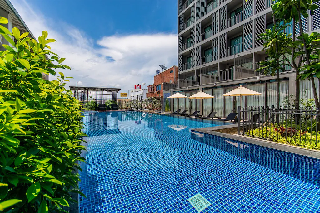 โรงแรมพัทยาติดทะเลแห่งนี้มีสระว่ายน้ำกลางแจ้งกระเบื้องสีน้ำเงินข้างอาคา ที่พักระยอง รโมเดิร์น มีเตียงอาบแดด ร่ม และต้นไม้ร่มรื่น ภายใต้ท้องฟ้าครึ้ม เหมาะกับวันพักผ่อนริมทะเล.