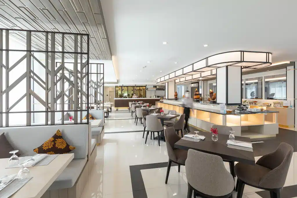 ร้านอาหารสมัยใหม่ภายในตกแต่งด้วยเฟอร์นิเจอร์โทนสีกลาง มีที่นั่งแบบมี  โรงแรมระยอง เบาะ ปูโต๊ะเรียบร้อย และพื้นที่บุฟเฟ่ต์ เหมาะสำหรับแขกที่พักในโรงแรมติดทะเลพัทยา ขณะที่พนักงานกำลังเดินใกล้เคาน์เตอร์.