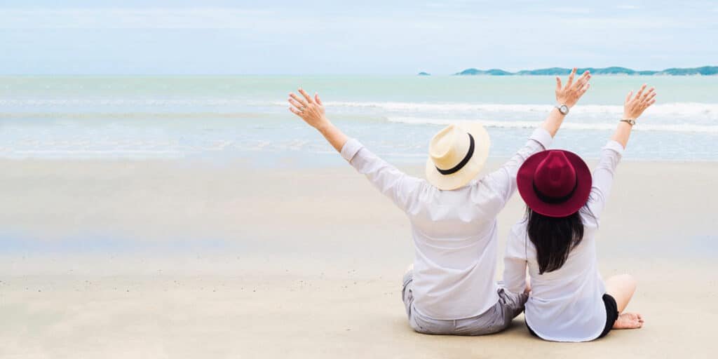 สองคนสวมหมวกนั่งอยู่บนชายหาดทราย หันหน้าไป ที่พักระยอง ทางทะเล ยกแขนขึ้น เพลิดเพลินกับวิวหน้า โรงแรมติดทะเลพัทยา.