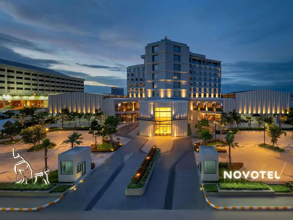โรงแรมทันสมัยพร้อมไฟส่องสว่าง ที่พักระยอง ภายนอกที่อบอุ่น ทางเข้าที่ตกแต่งด้วยภูมิทัศน์ และป้าย "Novotel" ที่ส่องสว่างในยามพลบค่ำ—โรงแรมพัทยาติดทะเลที่เหมาะสำหรับผู้ที่มองหาความสะดวกสบายใกล้ทะเล.