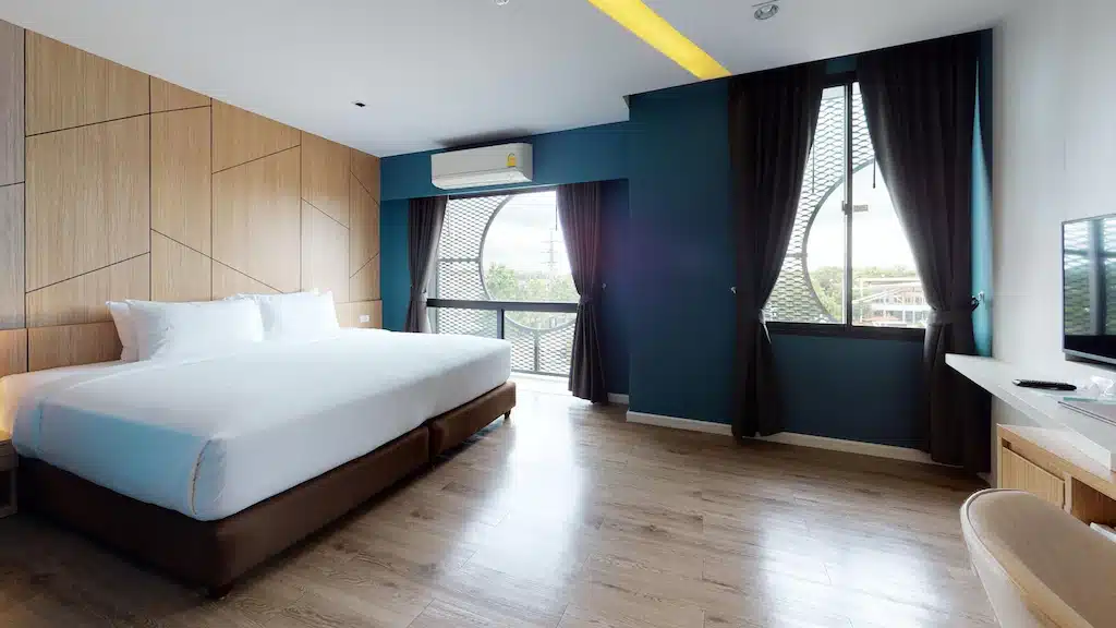ห้องพักโรงแรมสมัยใหม่พร้อมเตียงขนาดใหญ่ พื้นไม้ ผนังสีน้ำเงินเป็นจุดเด่น และหน้าต่างสองบานพร้อมผ้าม่านสีเข้ม—เหมาะสำหรับผู้ที่ต้องการ  ที่พักระยอง โรงแรมพัทยาติดทะเล มีทีวีติดผนัง โต๊ะทำงาน เก้าอี้ และเครื่องปรับอากาศ.