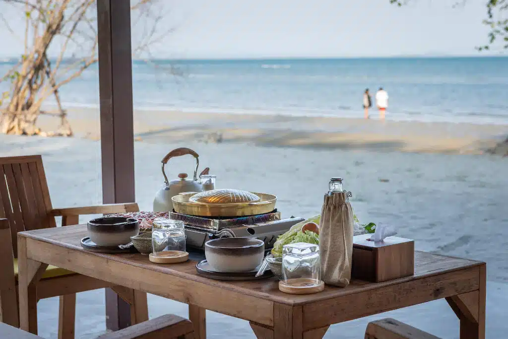 โต๊ะไม้ที่จัดวางอาหารและเครื่องดื่มไว้ มองเห็นชายหาดทรายที่ โรงแรมติดทะเลพัทยา โดยมีคนสองคนเดินอยู่ริมชายฝั่งเป็นฉากหลัง.  โรงแรมระยอง 