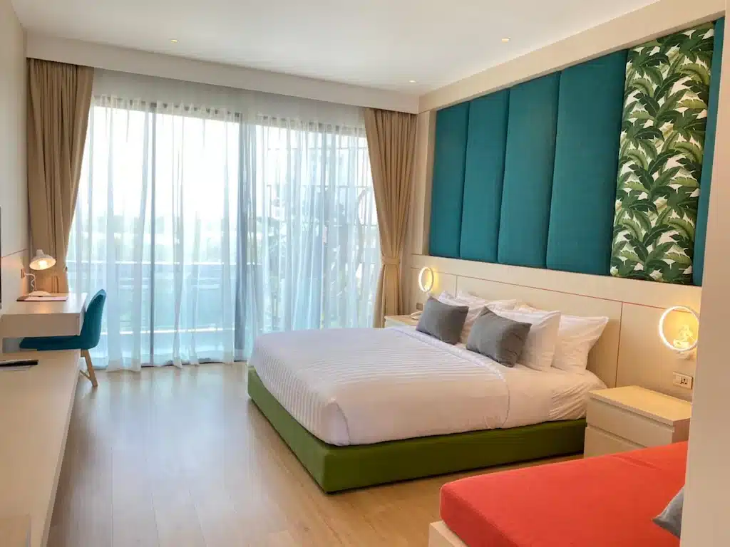 ห้องพักโรงแรมสมัยใหม่พร้อมเตียง ที่พักระยอง ขนาดใหญ่ หัวเตียงสีเขียวอมฟ้า แผงตกแต่งสไตล์ทรอปิคอล โต๊ะทำงาน ม้านั่งสีแดง และหน้าต่างบานใหญ่จากพื้นจรดเพดานพร้อมผ้าม่านบางและผ้าม่านสีเบจในโรงแรมติดทะเลพัทยาที่มีสไตล์.