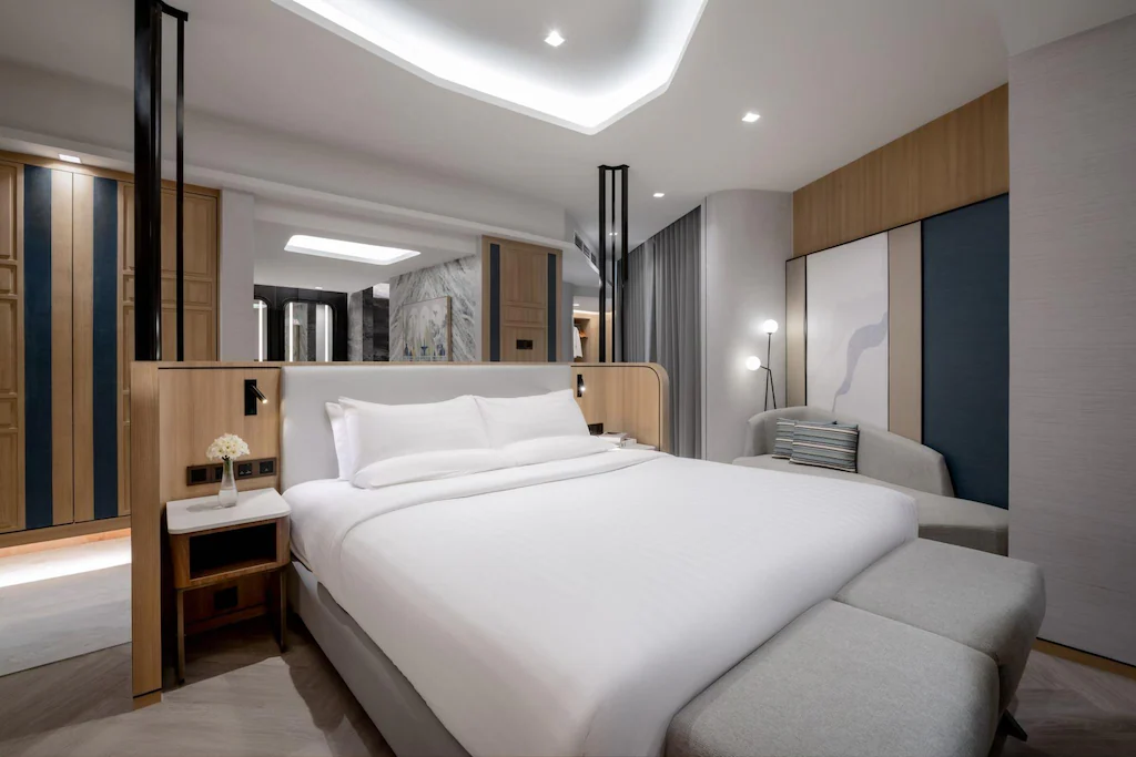 ห้องพักโรงแรมสมัยใหม่ที่ ที่พักติดทะเลพัทยา พร้อมเตียงขนาดใหญ่ที่จัดไว้อย่าง ที่พักระยอง เรียบร้อย ตกแต่งด้วยไม้ โซฟาสีเทา โต๊ะข้างเตียง และดีไซน์ร่วมสมัยที่สว่างไสว.