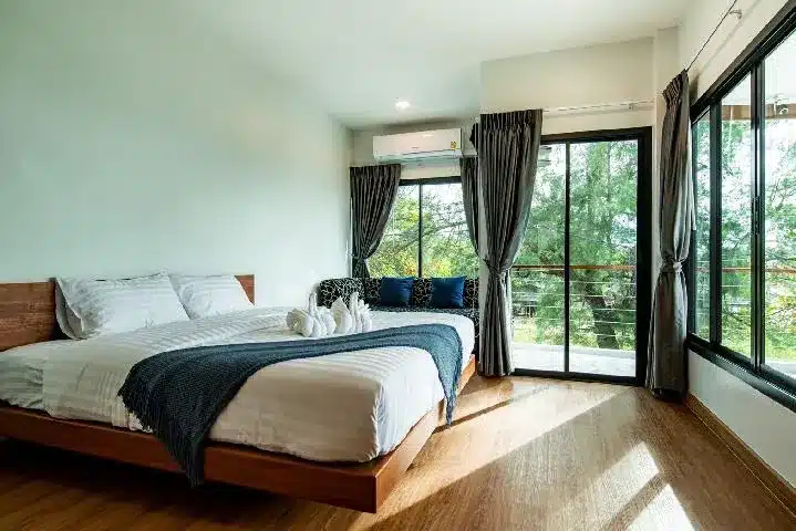 ห้องนอนสไตล์โมเดิร์นพร้อมเตียงขนาดใหญ่ ผ้าม่านสีเข้ม พื้นไม้ และหน้าต่างบานใหญ่จากพื้นจรดเพดานที่มองเห็นต้นไม้และระเบียงใน ที่พักพัทยาติดทะเล หรูหราสำหรับการพักผ่อนริมทะเลอย่างผ่อนคลาย.