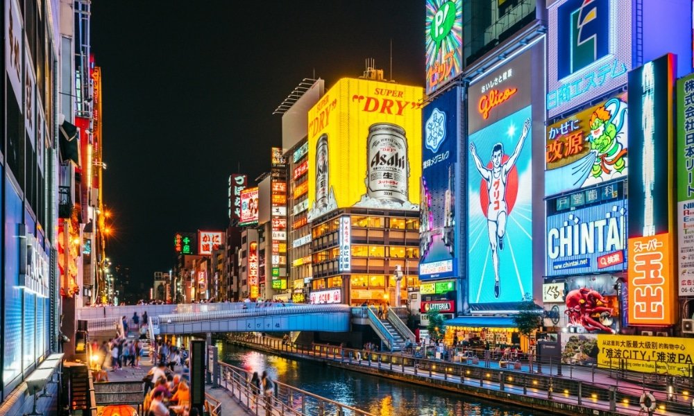 ถนนในเมืองที่คึกคักในยามค่ำคืนที่โอซาก้า ประเทศญี่ปุ่น มีป้ายไฟนีออนสว่างไสว คลอง และผู้คนเดินเล่นริมน้ำ—เหมาะสำหรับผู้ที่วางแผนจะเที่ยวญี่ปุ่นด้วยตัวเองและสำรวจสถานที่ท่องเที่ยวฮิตในญี่ปุ่น.