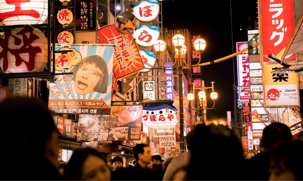 ถนนที่แออัดในเมืองญี่ปุ่นยามค่ำคืน รายล้อมไปด้วยป้ายไฟสว่างไสว ป้ายโฆษณา และโคมไฟที่มีตัวอักษรและภาพของญี่ปุ่น—เหมาะสำหรับผู้ที่ต้องการ เที่ยวญี่ปุ่นด้วยตัวเอง และค้นพบ สถานที่ท่องเที่ยวญี่ปุ่น ที่ไม่เหมือนใคร.