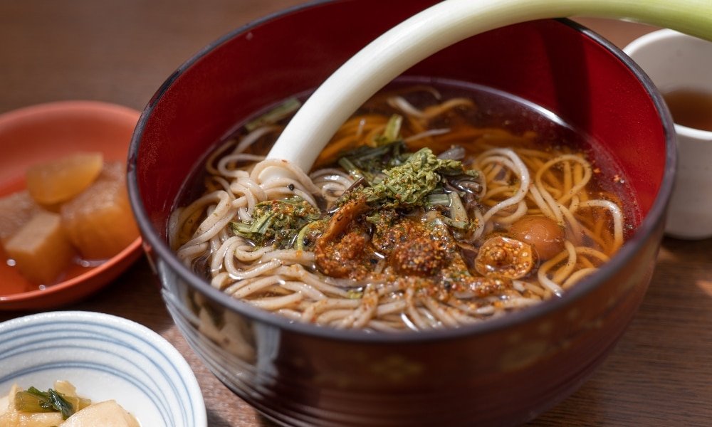 ชามบะหมี่โซบะร้อนเสิร์ฟพร้อมเครื่องเทศและต้นหอมญี่ปุ่นหนึ่งก้าน เสิร์ฟพร้อมเครื่องเคียงในชามเล็ก ๆ บนโต๊ะไม้—เหมาะสำหรับผู้ที่อยากเที่ยวญี่ปุ่นด้วยตัวเองและสัมผัสกับรสชาติญี่ปุ่นแท้ ๆ.
