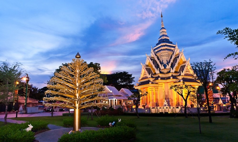 วัดที่สว่างไสวด้วยแสงไฟและสถาปัตยกรรมที่วิจิตรตั้งอยู่ข้างต้นไม้ประดับตกแต่งสีทองในยามพลบค่ำ ล้อมรอบด้วยพืชพรรณเขียวขจีและทางเดินที่ส่องสว่าง—เป็นหนึ่งในสถานที่ท่องเที่ยวที่ต้องไปเยือนในขอนแก่นสำหรับนักท่องเที่ยว.