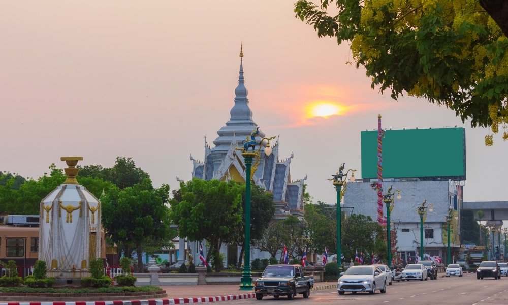 ถนนในเมืองขอนแก่นที่มีรถยนต์, ต้นเสาไฟสีเขียว, และอาคารสีเงินคล้ายวัดอยู่ใต้ท้องฟ้าที่มีแสงอาทิตย์ตกดินเป็นหมอก—เหมาะสำหรับผู้ที่กำลังมองหาที่เที่ยวในขอนแก่น. ต้นไม้และป้ายโฆษณาขนาดใหญ่ที่ว่างเปล่าทำให้ภาพนี้สมบูรณ์.