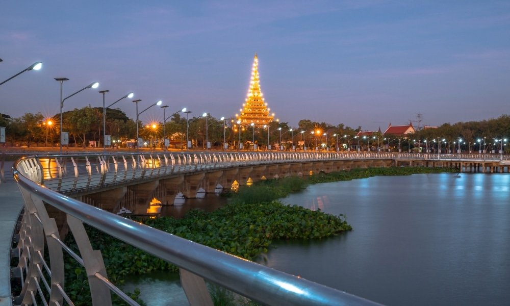สะพานคนเดินสมัยใหม่ที่มีราวบันไดทอดข้ามน้ำไปยังโครงสร้างวัดที่มีแสงไฟสว่างไสวเป็นชั้น ๆ ในยามพลบค่ำ—เป็นสถานที่ท่องเที่ยวที่เป็นสัญลักษณ์ของขอนแก่นที่เหมาะสำหรับผู้ที่ต้องการสำรวจสถานที่ท่องเที่ยวที่น่าจดจำในขอนแก่น.