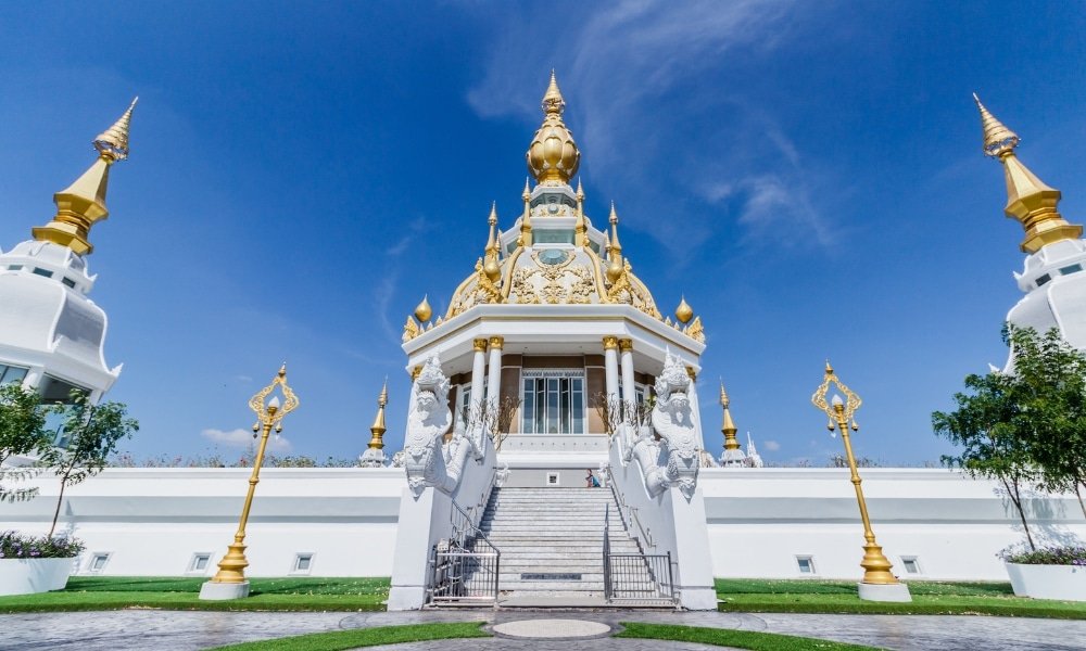วัดสีขาวและทองที่มียอดแหลมประดับประดาอย่างวิจิตร การออกแบบที่สมมาตร และองค์ประกอบตกแต่ง ตั้งอยู่ท่ามกลางท้องฟ้าสีฟ้าใส—หนึ่งในสถานที่ท่องเที่ยวที่ต้องไปเยือนในขอนแก่นสำหรับนักท่องเที่ยวที่สนใจวัฒนธรรม.