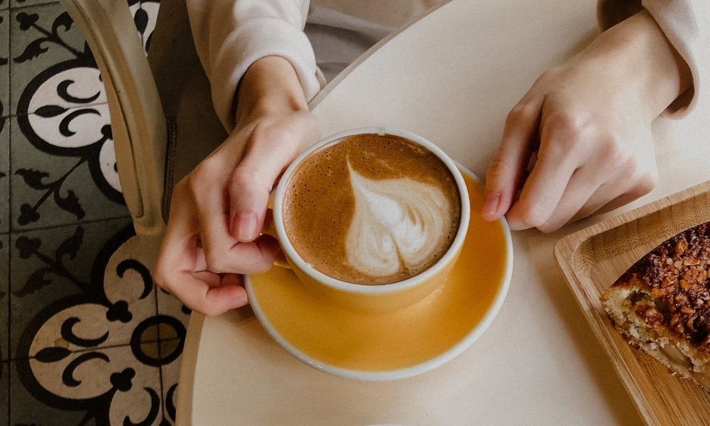 คนคนหนึ่งกำลังเพลิดเพลินกับกาแฟถ้วยสีเหลืองที่มีลาเต้อาร์ตเป็นรูปหัวใจบนโต๊ะกลม ข้างๆ มีจานไม้ที่มีขนมอบ—เหมาะสำหรับการผ่อนคลายหลังจากเที่ยวชม ขอนแก่นที่เที่ยว.