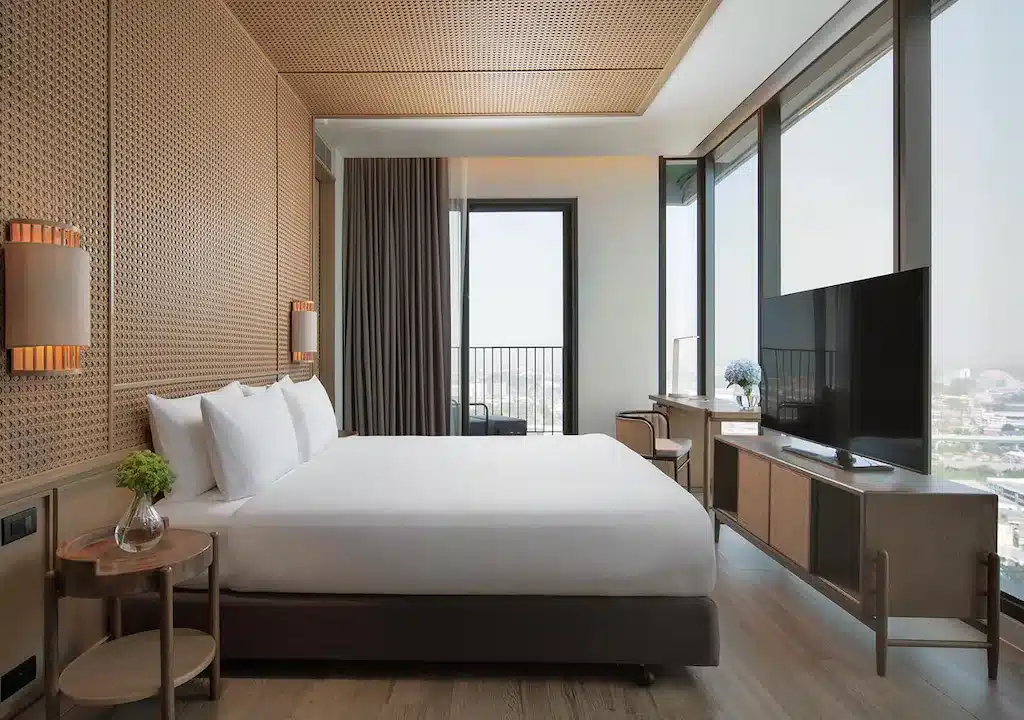 ห้องพักโรงแรมสมัยใหม่พร้อมเตียงขนาดใหญ่ ทีวีติดผนัง โต๊ะข้างพร้อมต้นไม้ และหน้าต่างบานใหญ่จากพื้นจรดเพดานที่มองเห็นวิวเมือง—เหมาะสำหรับการพักผ่อนหลังเที่ยวขอนแก่นและสำรวจสถานที่ท่องเที่ยวยอดนิยมในขอนแก่น.