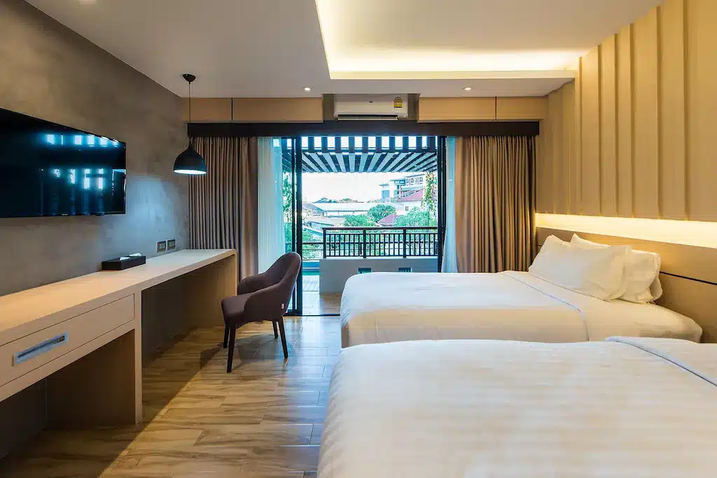 ห้องพักโรงแรมสมัยใหม่พร้อมเตียงคู่สองเตียง โทรทัศน์ติดผนัง พื้นไม้ และระเบียงพร้อมวิว—เหมาะสำหรับการพักผ่อนหลังจากสำรวจสถานที่ท่องเที่ยวในขอนแก่นระหว่างการผจญภัยท่องเที่ยวขอนแก่นของคุณ.