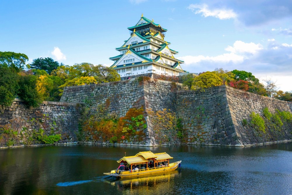 เรือลำสีเหลืองแบบดั้งเดิมลอยอยู่ในคูน้ำหน้าปราสาทโอซาก้า ล้อมรอบด้วยกำแพงหินและต้นไม้ในฤดูใบไม้ร่วงภายใต้ท้องฟ้าที่มีเมฆบางส่วน—เหมาะสำหรับผู้ที่กำลังวางแผนท่องเที่ยวญี่ปุ่นด้วยตัวเอง.